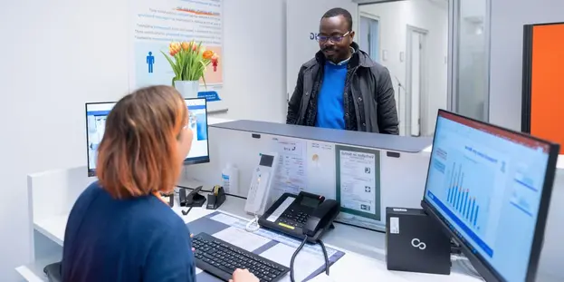 A man is standing at the reception desk of the Prevention Outpatient Clinic, talking to a staff member at a desk with two monitors.