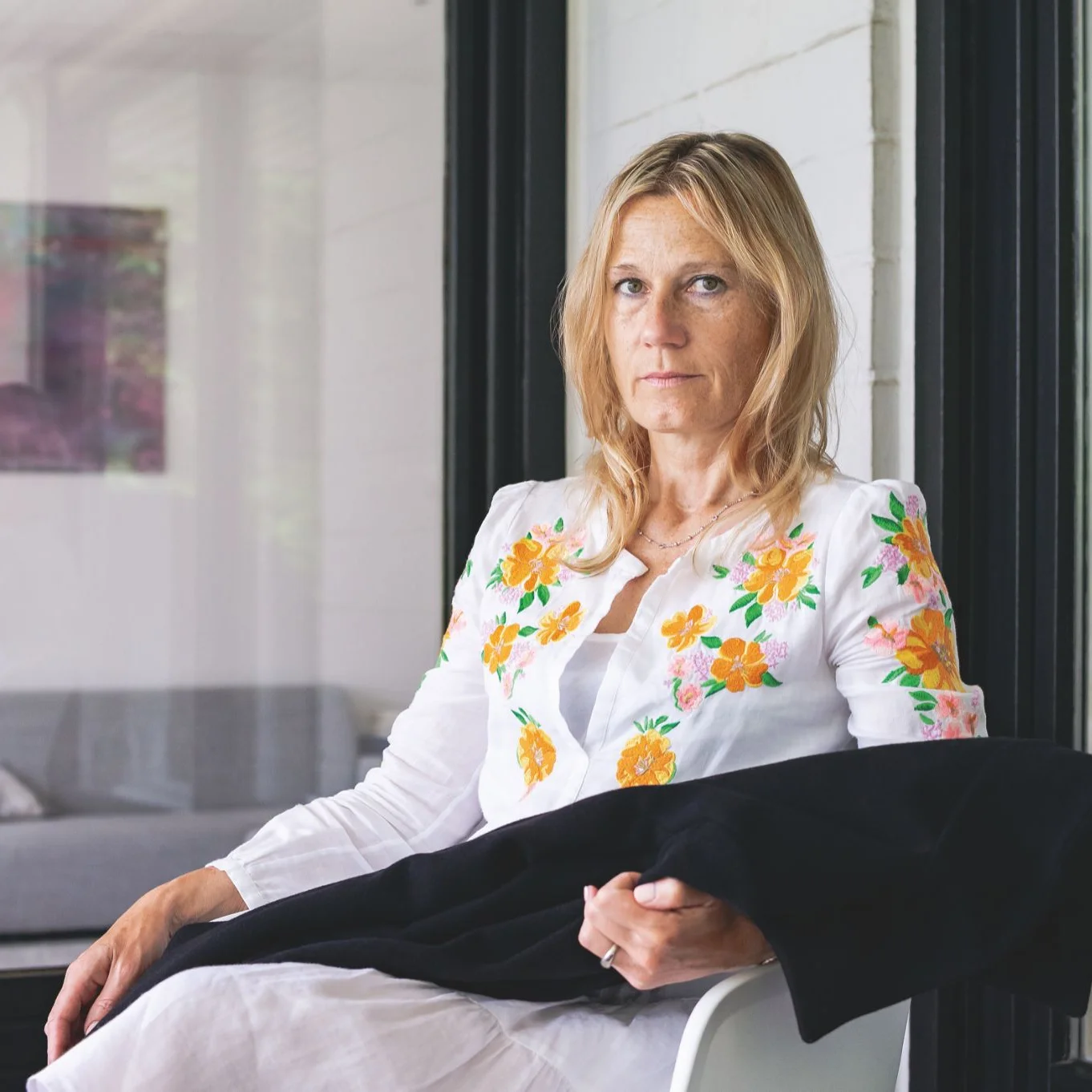 Frau mit blondem Haar und weißem Kleid mit Blumenmuster sitzt auf einem Stuhl und blickt ernst in die Kamera.
