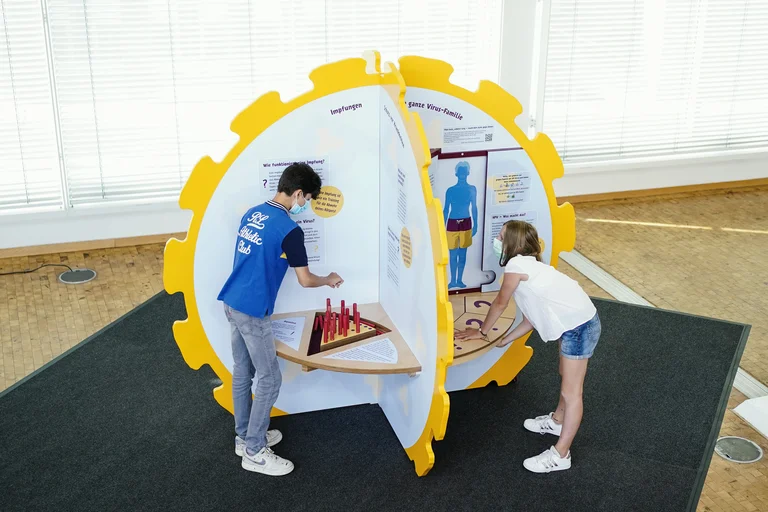 Zwei Kinder stehen vorgebeugt an der HPV-Mitmachstation, die als großes rundes HP-Virus dargestellt ist.
