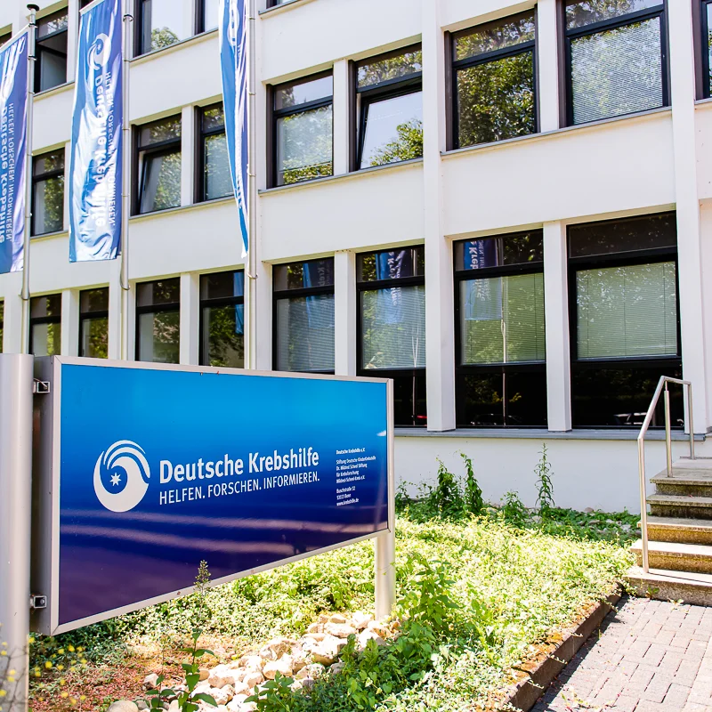  Außenansicht der Geschäftsstelle der Deutschen Krebshilfe mit blauem Firmenschild und Fahnen.