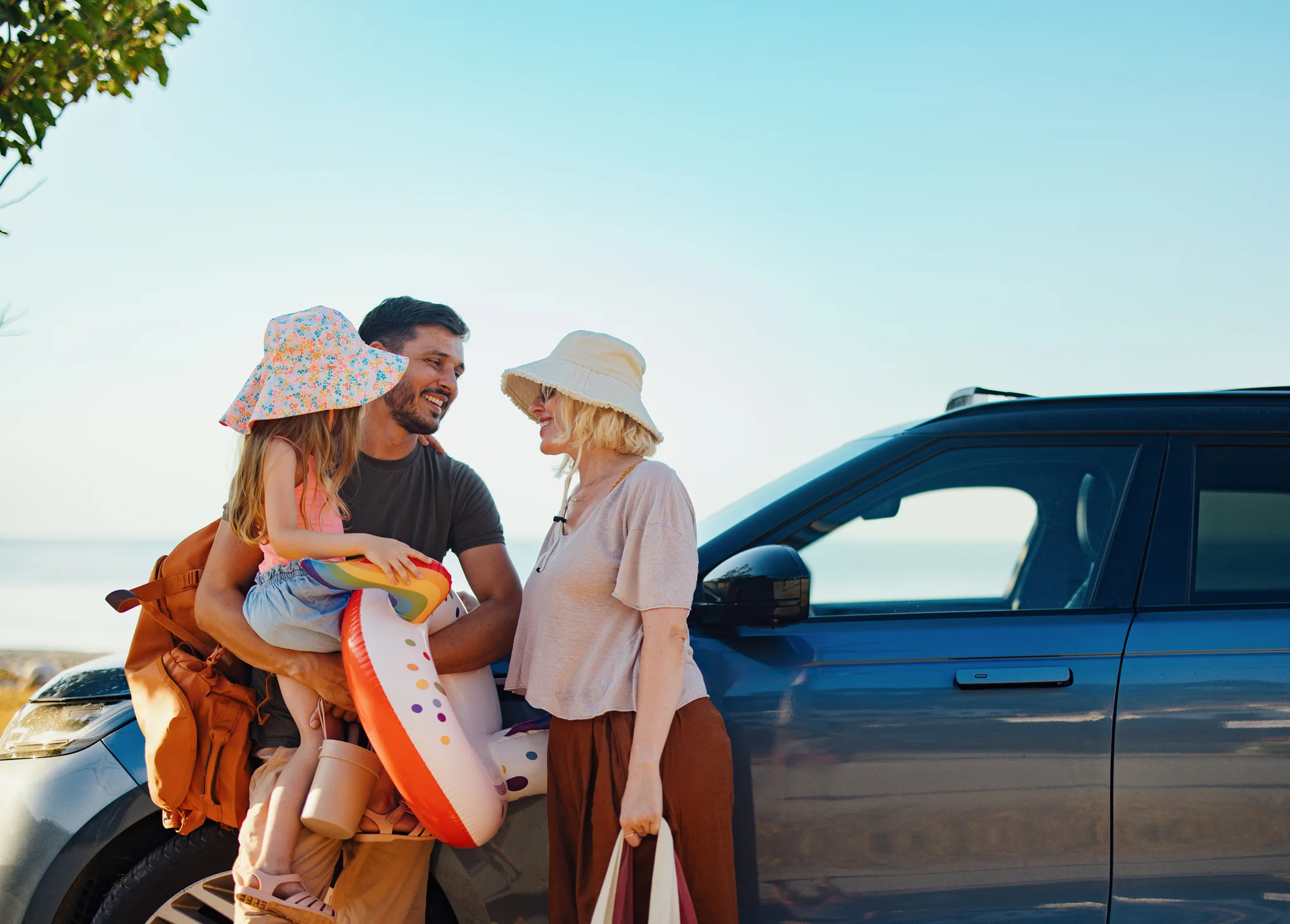 Eine dreiköpfige Familie mit Hüten und Strandspielzeug steht lächelnd an einem blauen Auto vor einer Meereskulisse.