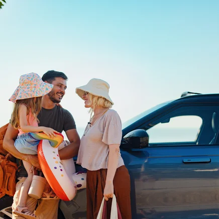 Eine dreiköpfige Familie mit Hüten und Strandspielzeug steht lächelnd an einem blauen Auto vor einer Meereskulisse.