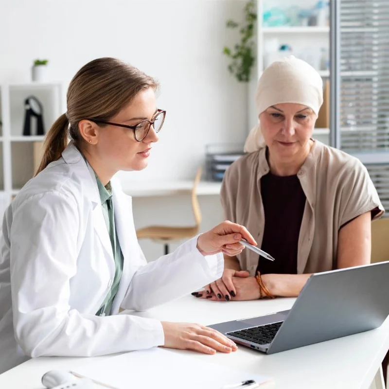 A doctor talks to a cancer patient wearing a head covering and points to something on the laptop screen.
