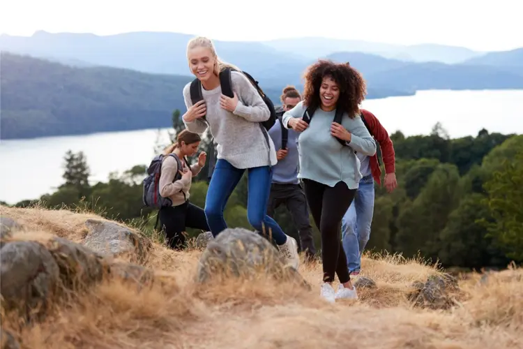 Gruppe junger Menschen wandert lachend durch hügelige Natur mit See im Hintergrund.