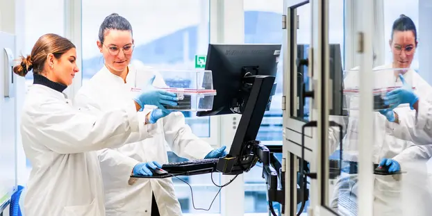 Forscherinnen des Deutschen Krebsforschungszentrums. Eine Frau steht vor einem Bildschirm und betrachtet eine durchsichtige Box, die eine andere Frau in den Händen hält. 