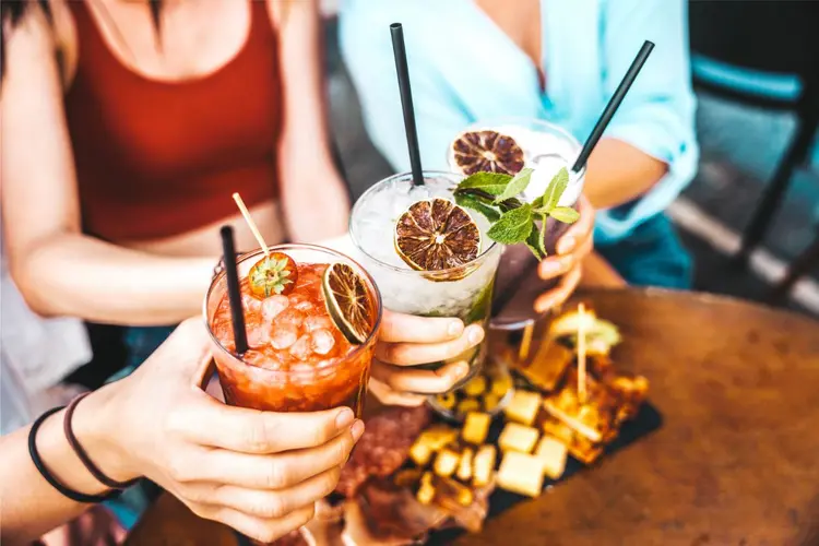Drei Personen stoßen mit bunten Cocktails an, im Hintergrund eine Platte mit Käse und Wurst.