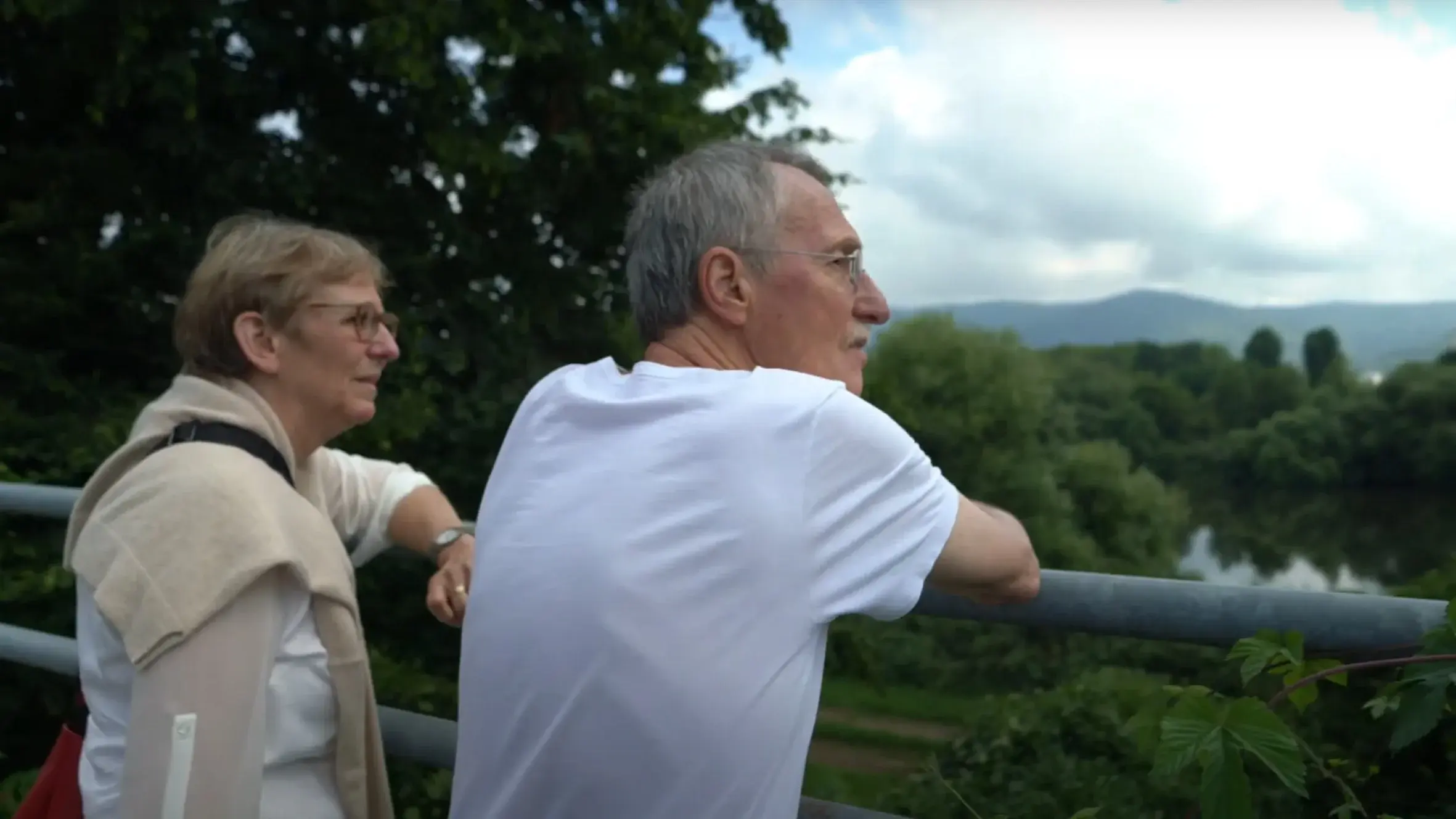 Zwei ältere Personen lehnen an einem Geländer und blicken in eine grüne Landschaft.